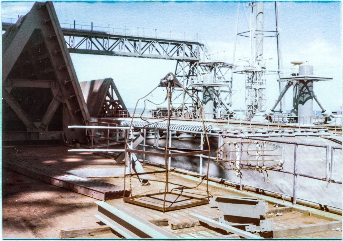 Image 012. Up on the pad deck at Space Shuttle Launch Complex 39-B, Kennedy Space Center, Florida. You are looking northeast, across the Flame Trench with the Flame Deflector topped by its row of Spray Headers filling the view of the Trench, and just left of that, on the far Trench wall, the comb-like row of nozzles on another Spray Header. At far left, shadowed by the RSS and FSS which both loom far above you into the sky just out of frame farther left, one of the large triangular-shaped Side Flame Deflectors sits parked in its Launch Position, and across the Trench in partial sunlight its mate sits in its Park Position. To the right of that, a confused tangle of steel comprises the North Piping Bridge, LH2 Tower, MLP Mount Pedestals, Sound Suppression System Water Tower, SSW supply risers for the MLP, and the Pneumatics Tower, not in that order, but before this is over, you will know them all, well. Center of frame, resting on the pad deck among pieces of loose steel and equipment, the Skip Box which you would ride, lifted by the crane to otherwise inaccessible locations on the tower, sits at the ready. To its right, secured to the removable handrail that guards the five story vertical drop to the bottom of the Flame Trench just beyond it, a stretcher can be seen, also at the ready, in case someone needed evacuation following serious injury. Photo by James MacLaren. Image 012. Up on the pad deck at Space Shuttle Launch Complex 39-B, Kennedy Space Center, Florida. You are looking northeast, across the Flame Trench with the Flame Deflector topped by its row of Spray Headers filling the view of the Trench, and just left of that, on the far Trench wall, the comb-like row of nozzles on another Spray Header. At far left, shadowed by the RSS and FSS which both loom far above you into the sky just out of frame farther left, one of the large triangular-shaped Side Flame Deflectors sits parked in its Launch Position, and across the Trench in partial sunlight its mate sits in its Park Position. To the right of that, a confused tangle of steel comprises the North Piping Bridge, LH2 Tower, MLP Mount Pedestals, Sound Suppression System Water Tower, SSW supply risers for the MLP, and the Pneumatics Tower, not in that order, but before this is over, you will know them all, well. Center of frame, resting on the pad deck among pieces of loose steel and equipment, the Skip Box which you would ride, lifted by the crane to otherwise inaccessible locations on the tower, sits at the ready. To its right, secured to the removable handrail that guards the five story vertical drop to the bottom of the Flame Trench just beyond it, a stretcher can be seen, also at the ready, in case someone needed evacuation following serious injury. Photo by James MacLaren.